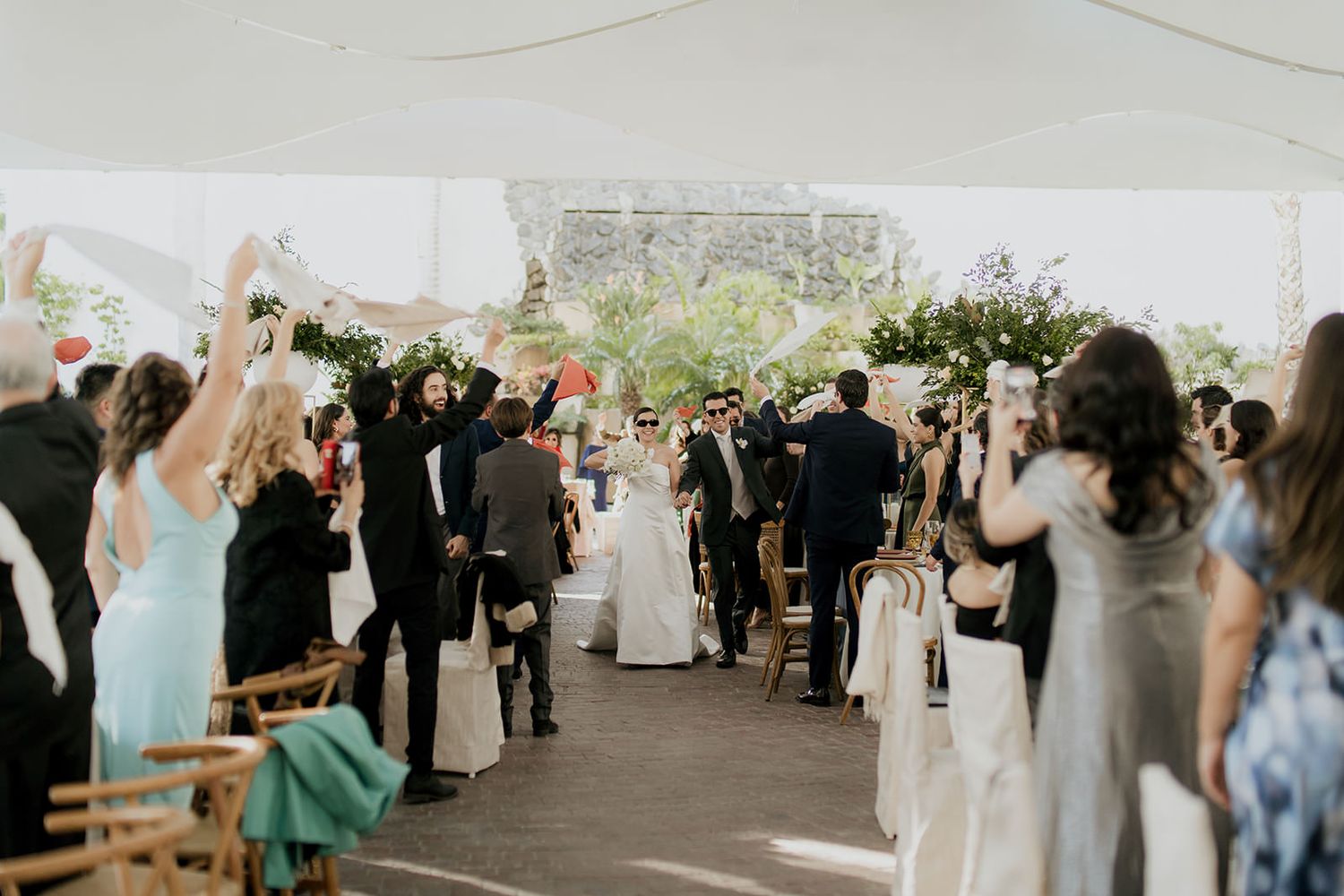Newlyweds walk down the aisle as cheering guests celebrate at an outdoor wedding ceremony.