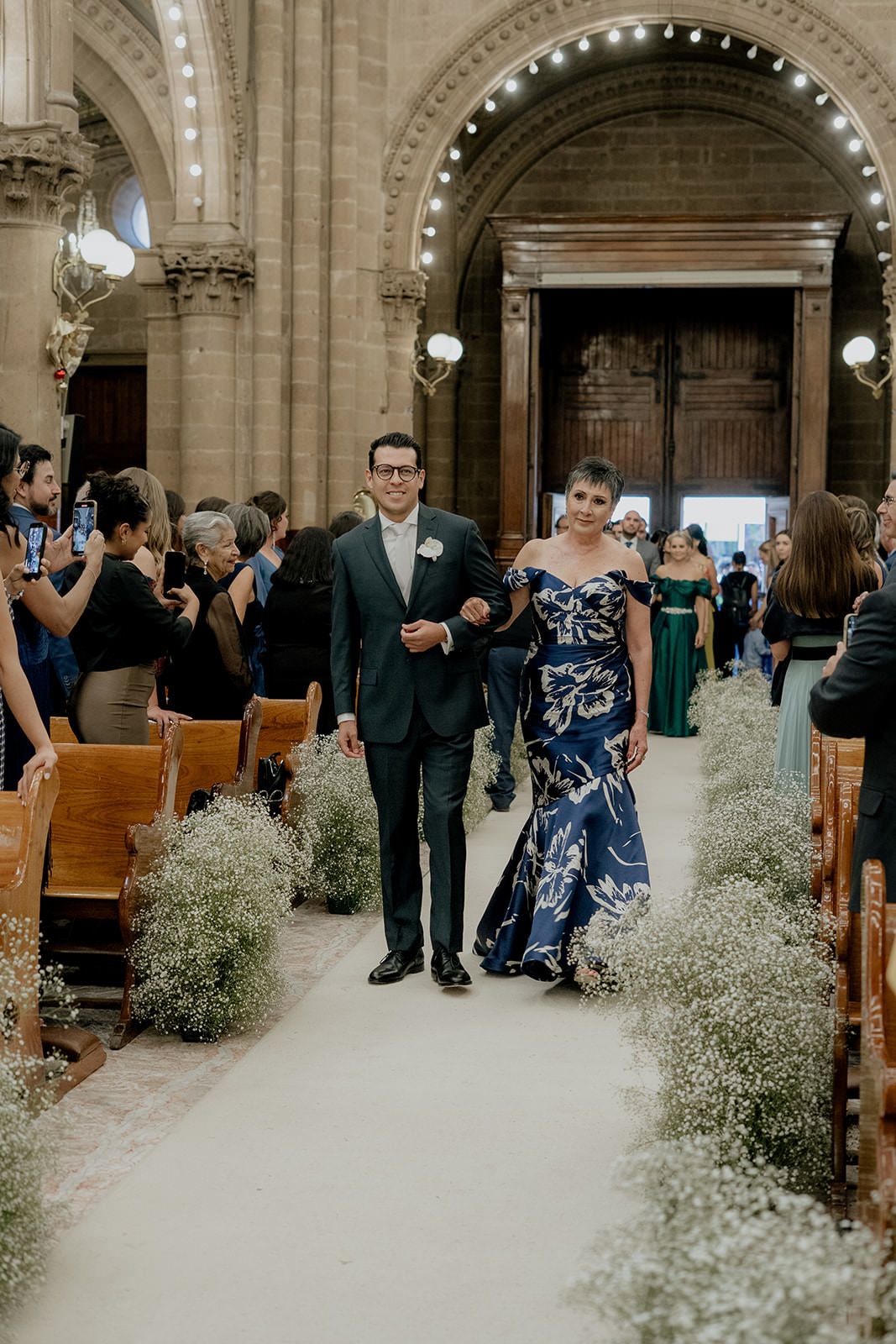 Man in dark suit escorts woman in blue floral gown down church aisle lined with baby's breath at wedding ceremony.