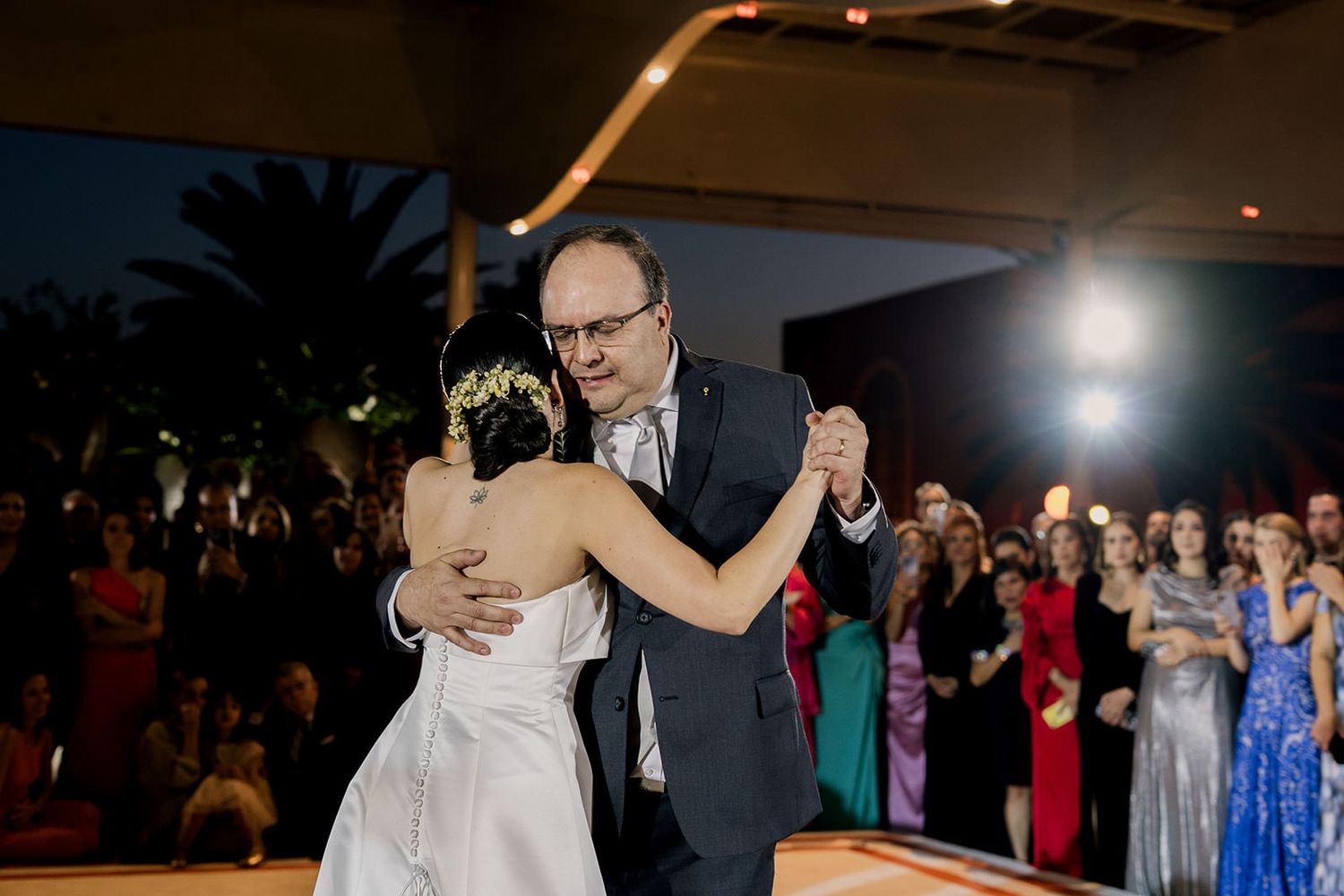 Bride dancing with older man, likely father, during emotional wedding reception dance at night.