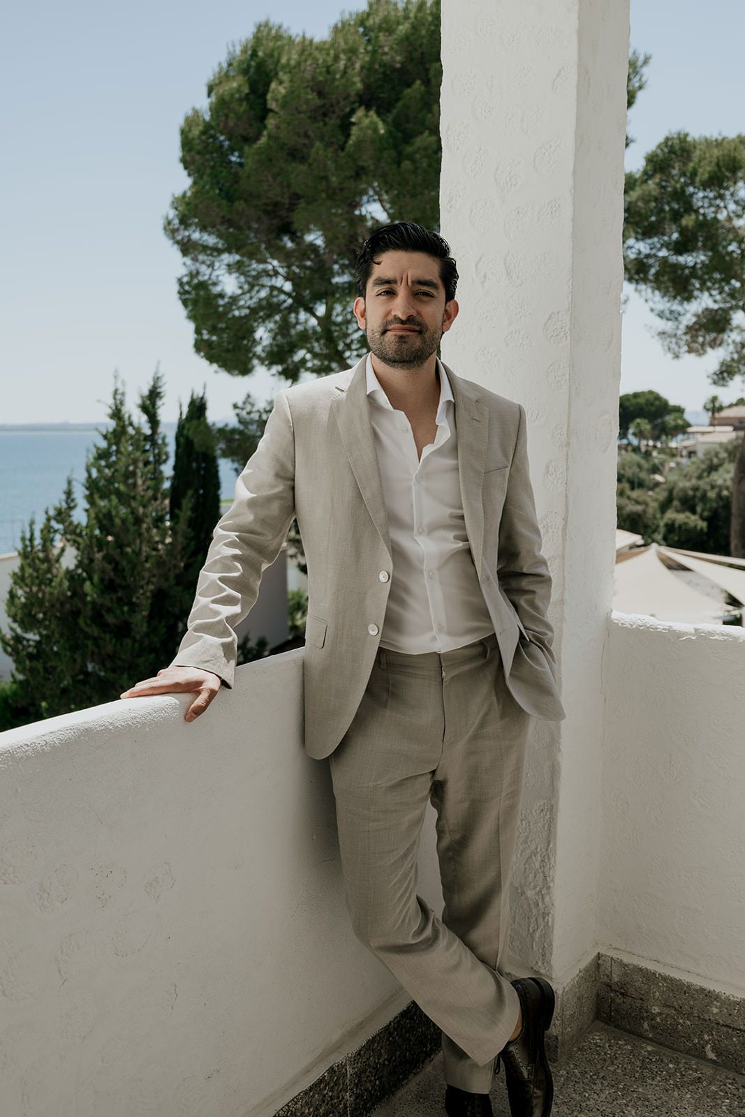 Elegant beige suit against white Mediterranean architecture with scenic backdrop.