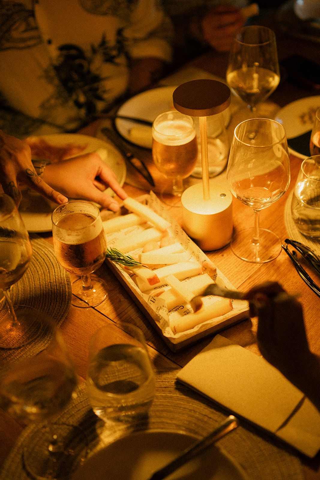 Hands reach for wine glasses and appetizers on a wooden table illuminated by warm candlelight in a cozy setting.
