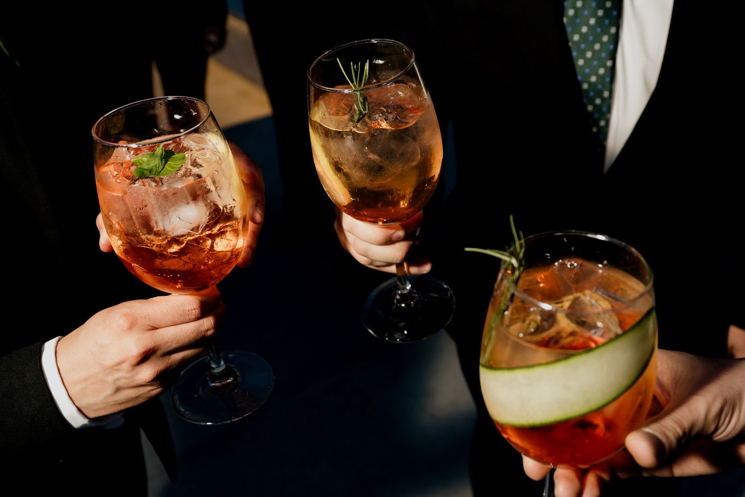 Three guests clink colorful cocktails garnished with herbs and fruit at wedding reception.