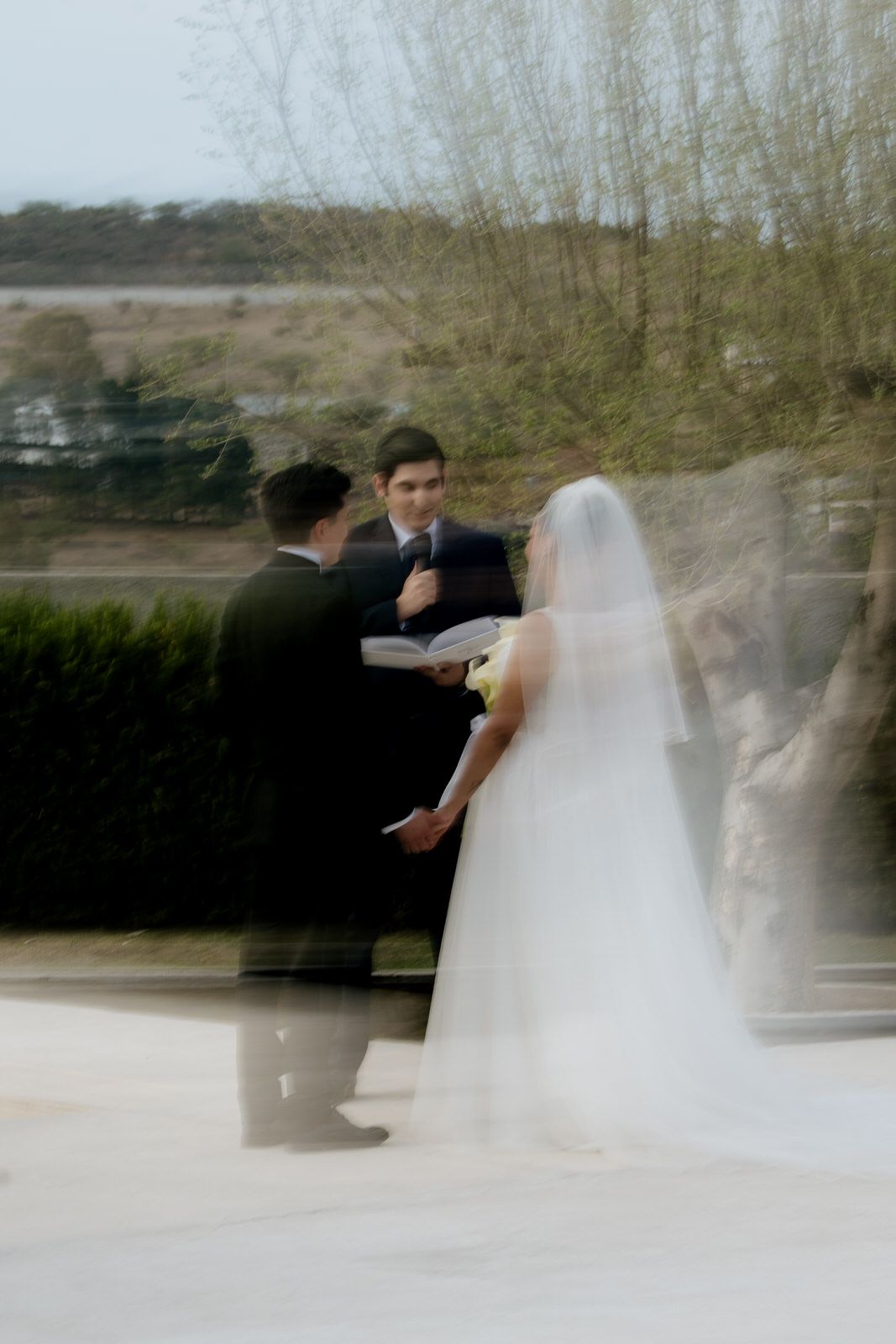 Blurred artistic shot of bride in white veil and groom during outdoor wedding ceremony with officiant.