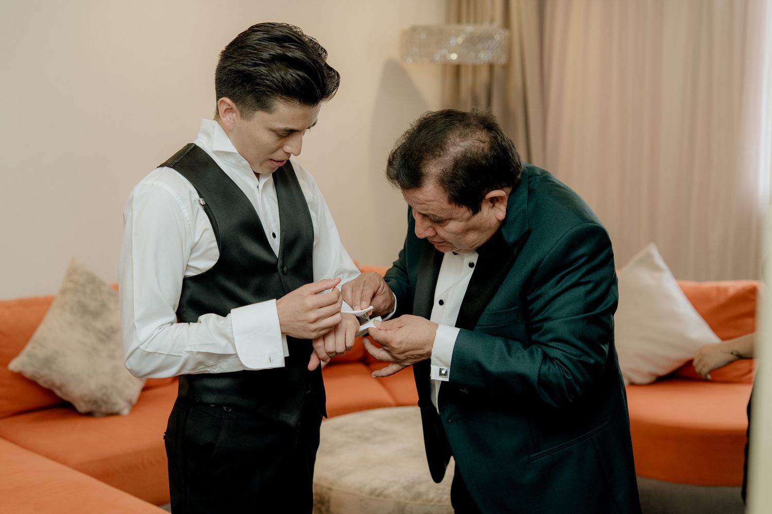 Father helping groom with cufflinks while getting ready for wedding ceremony in elegant hotel room.