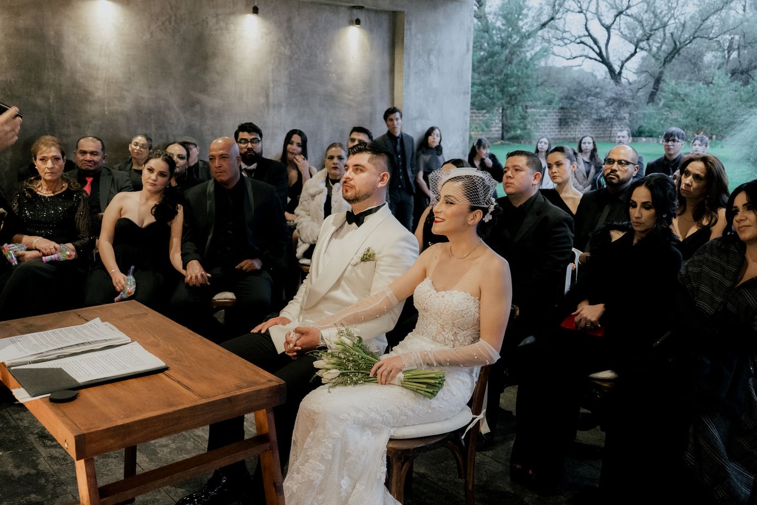 A couple sits at a rustic wooden table during their wedding ceremony with guests in black attire.