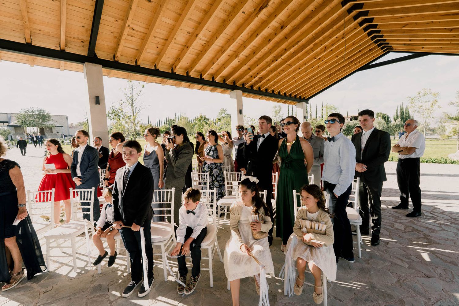 Wedding guests fill covered outdoor venue with wooden ceiling during ceremony proceedings.