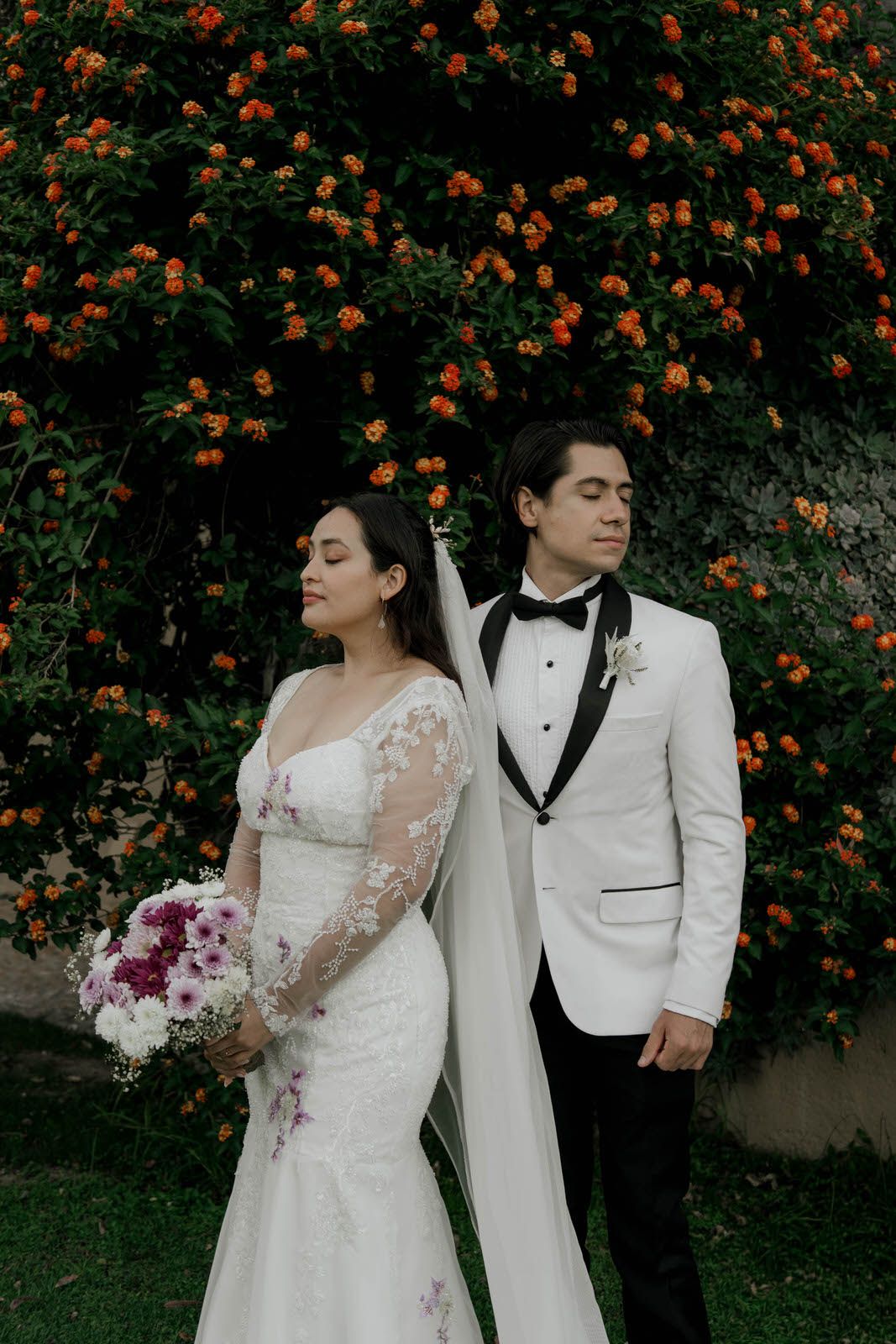Elegant wedding couple poses against vibrant orange flowering bush in garden setting.
