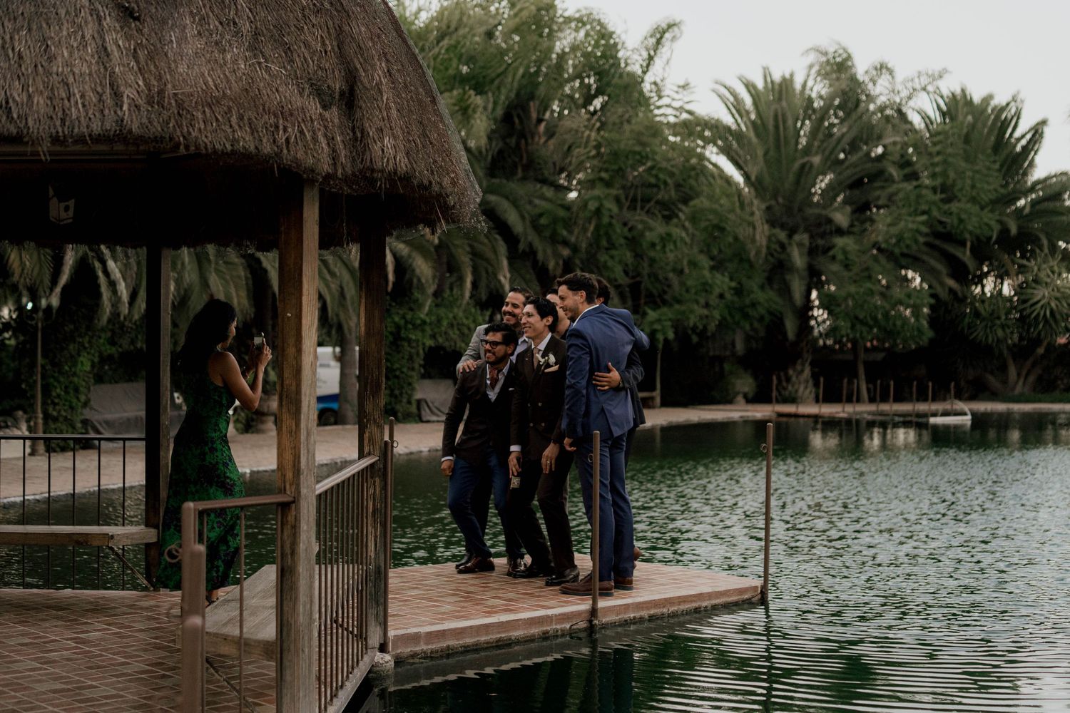 Wedding party gathered on a wooden dock surrounded by palm trees and water.