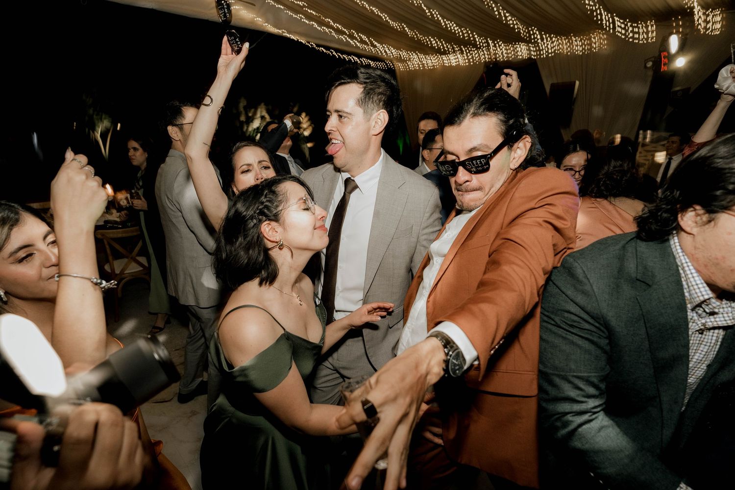 Lively wedding reception crowd dances beneath romantic string lighting and fabric draping.