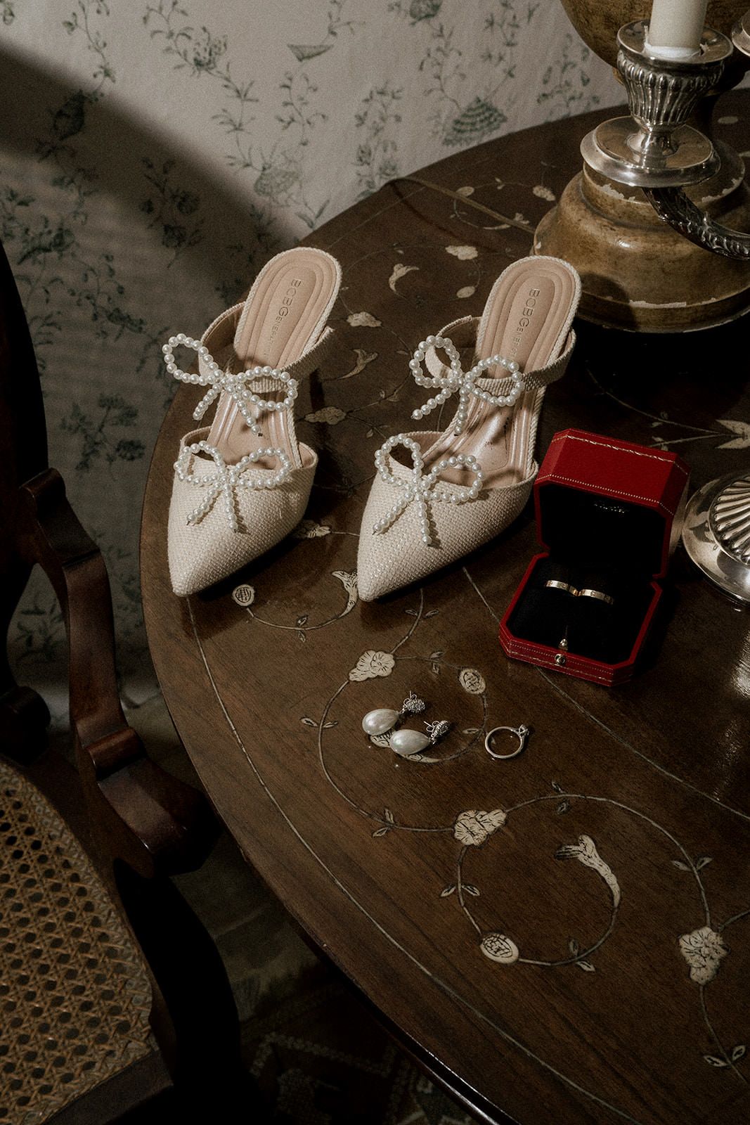 Elegant white bridal shoes and wedding rings displayed on a dark wooden table with decorative details.