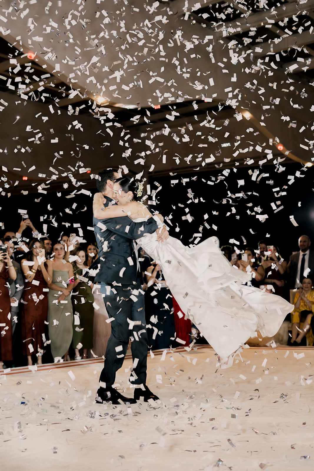 Bride and groom share first dance surrounded by falling white confetti at elegant wedding reception.