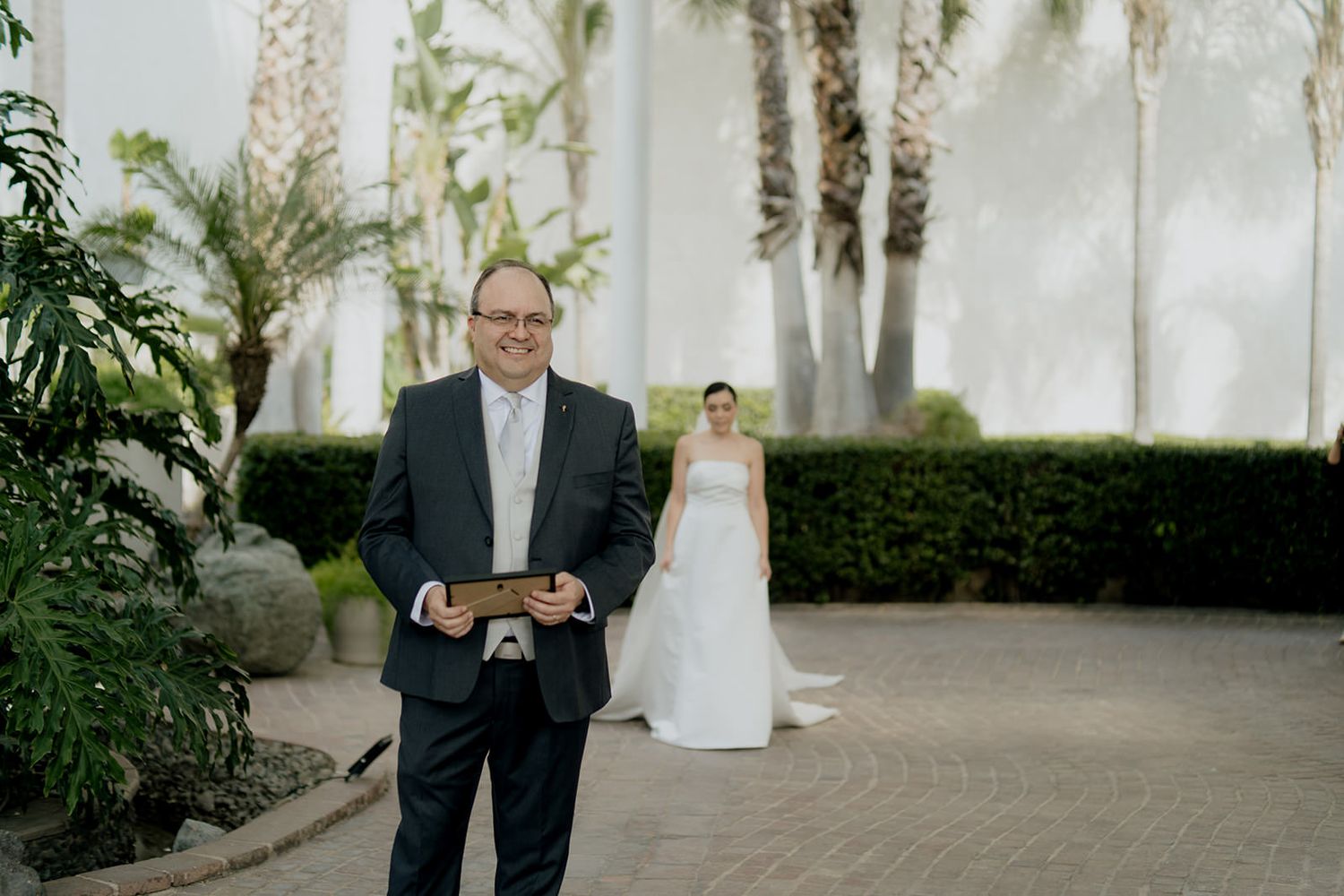 Groom holding a book smiles as bride approaches in a stunning white gown at outdoor wedding venue.