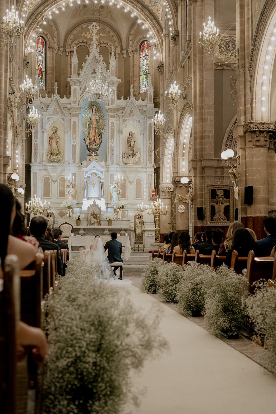 Bride and groom kneel at ornate church altar during wedding ceremony, flanked by floral arrangements and wooden pews.