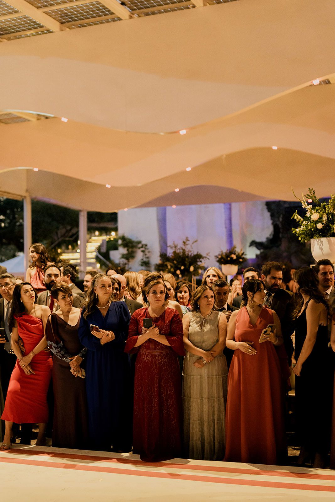 Elegantly dressed wedding guests gather under a warmly lit outdoor tent during an evening reception celebration.