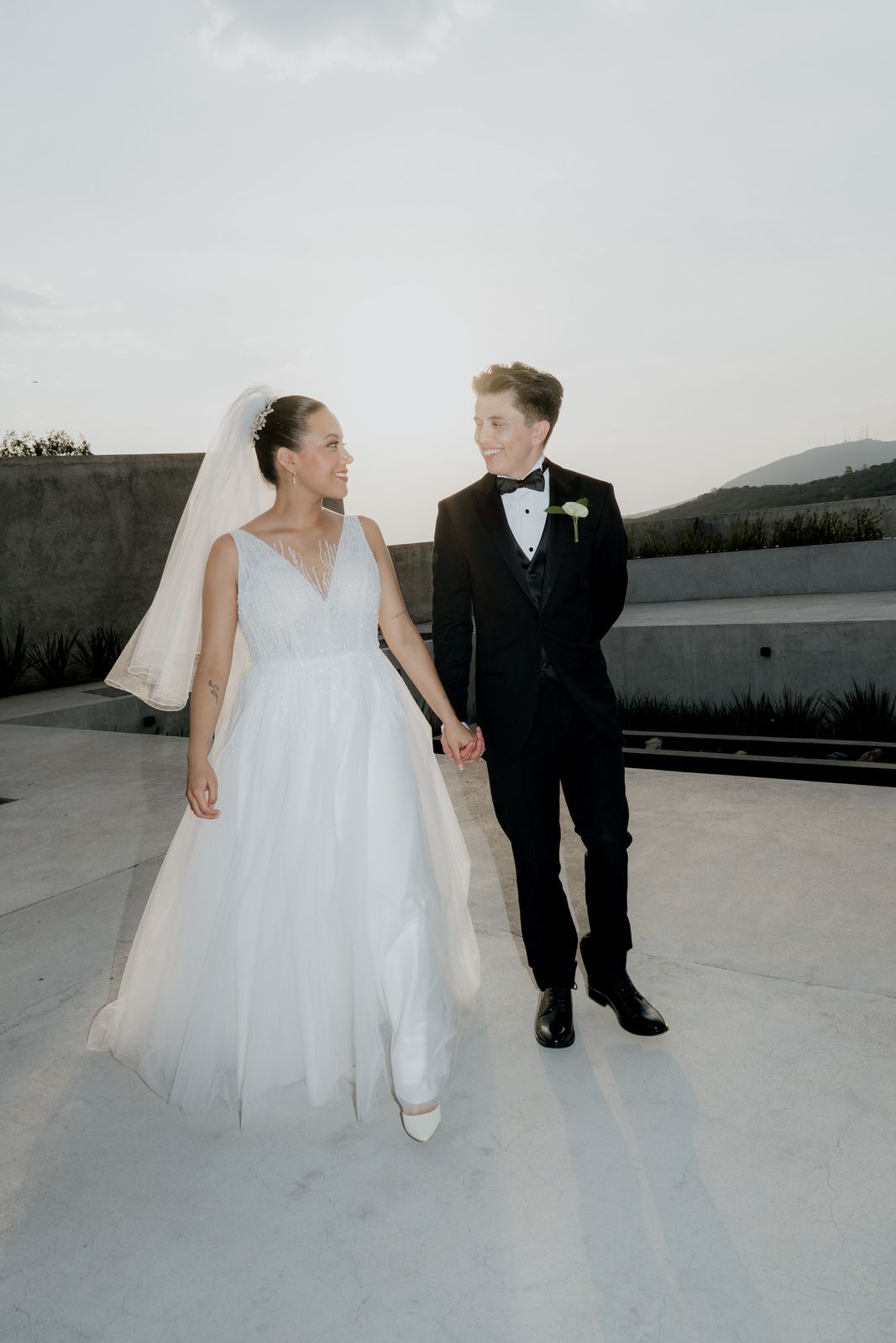 Newlyweds hold hands on rooftop terrace at dusk, bride in tulle gown and groom in black tuxedo gazing at each other.