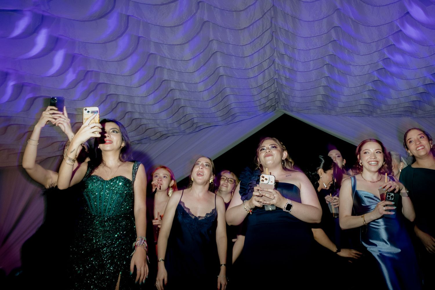 Group of women in formal gowns taking selfies and celebrating at an elegant evening event under decorative ceiling.