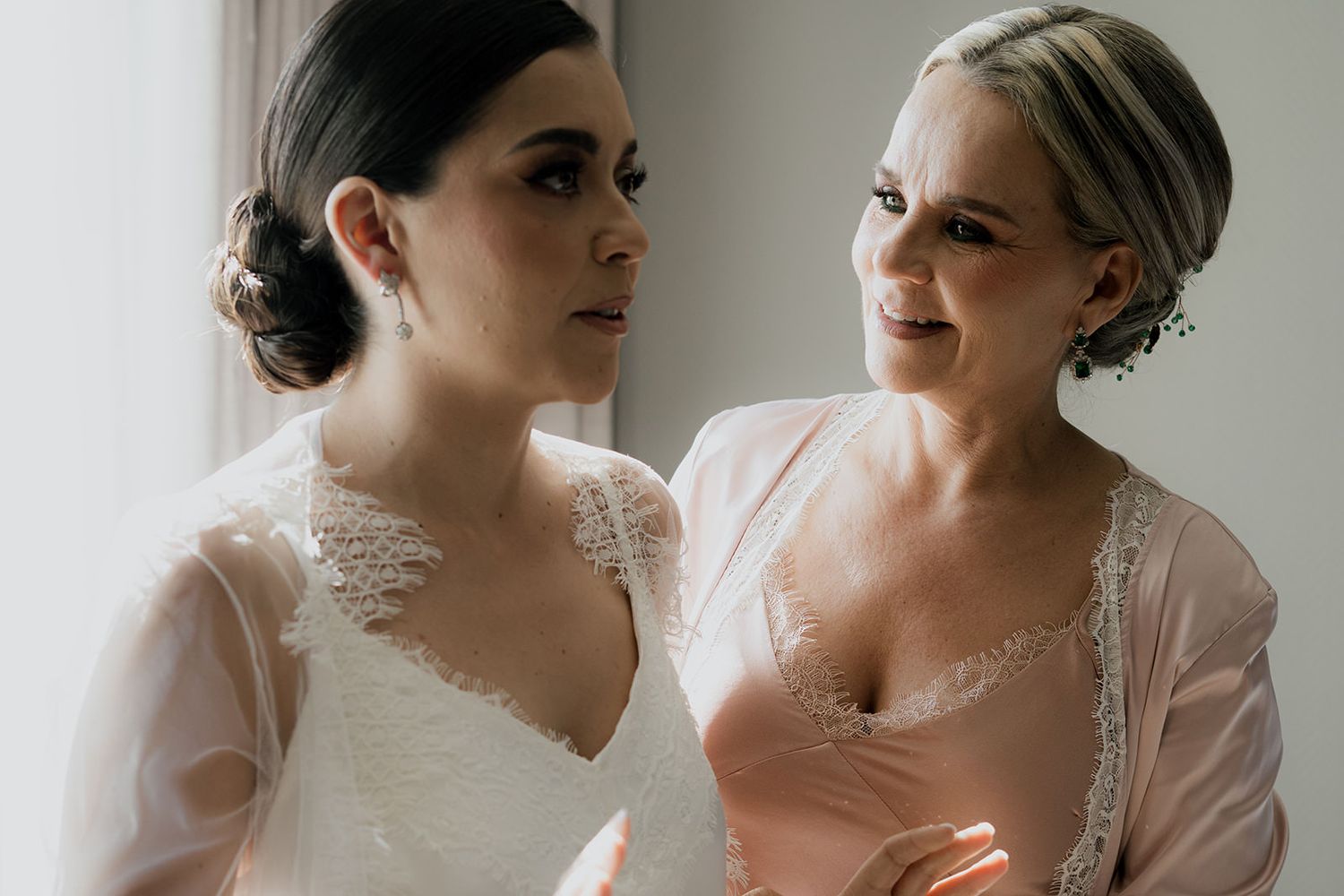 Bride in white lace wedding dress with bridesmaid in pink robe during wedding preparations.