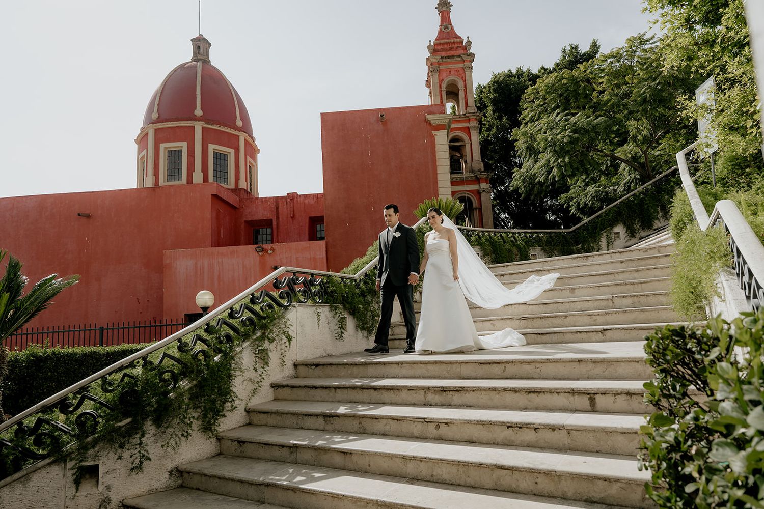 Bride and groom walk down stone steps at a stunning Mexican wedding venue with red domed church and bell tower backdrop.