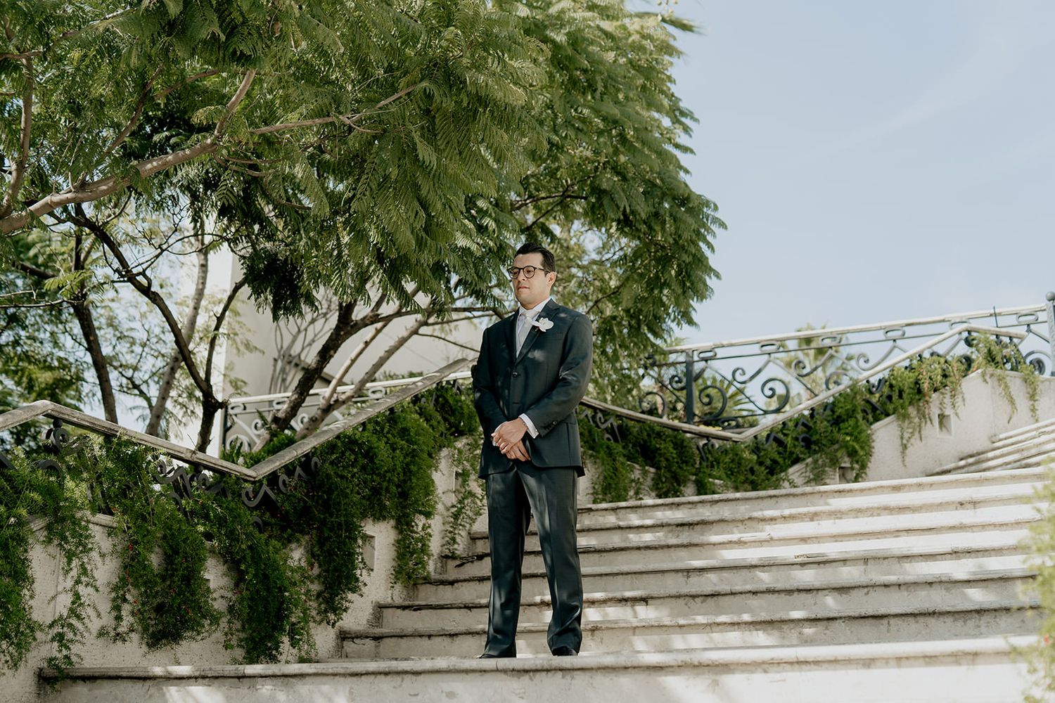 Dapper groom in dark suit standing on ivy-covered stone staircase at outdoor wedding venue.