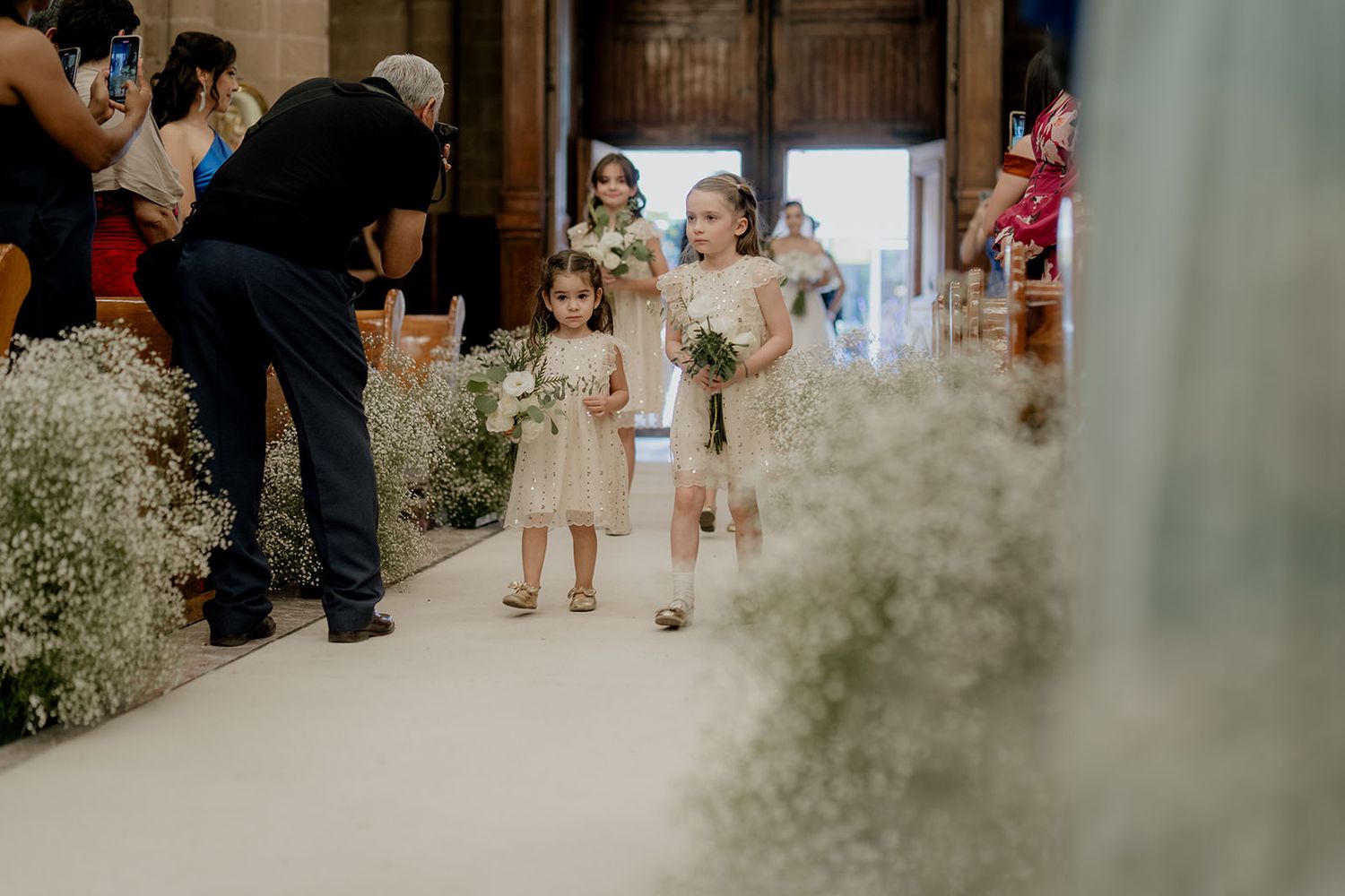 Two flower girls in gold dresses walk down a white wedding aisle adorned with white floral arrangements.