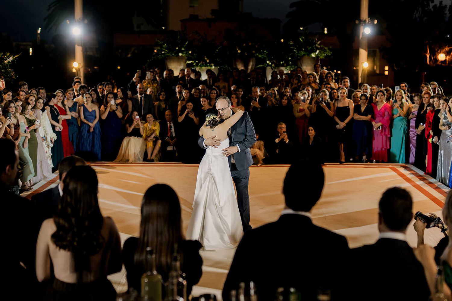 Bride and groom share first dance surrounded by elegantly dressed wedding guests at an outdoor evening reception.