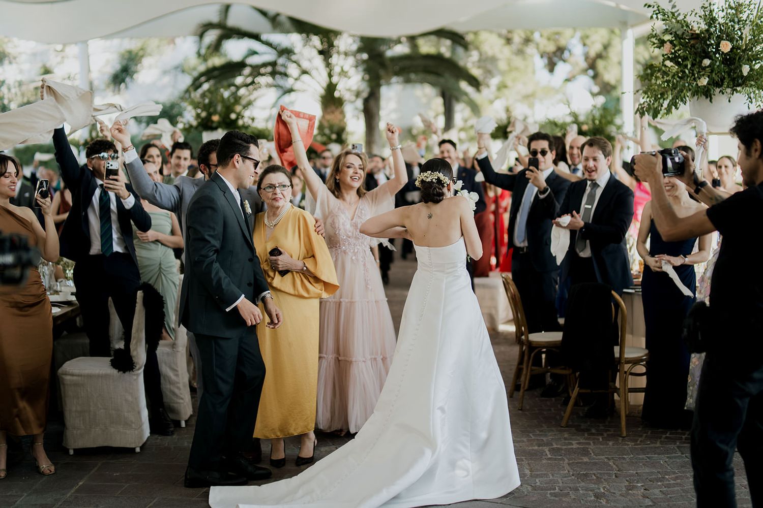 Bride and groom walk down the aisle as wedding guests celebrate and cheer at an outdoor reception.