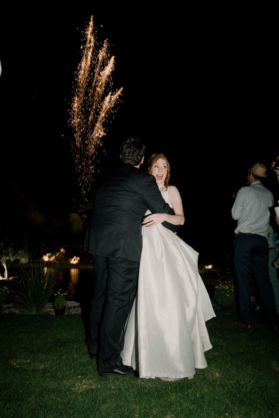 Sparklers illuminate the night as wedding guests gather on a grassy lawn.