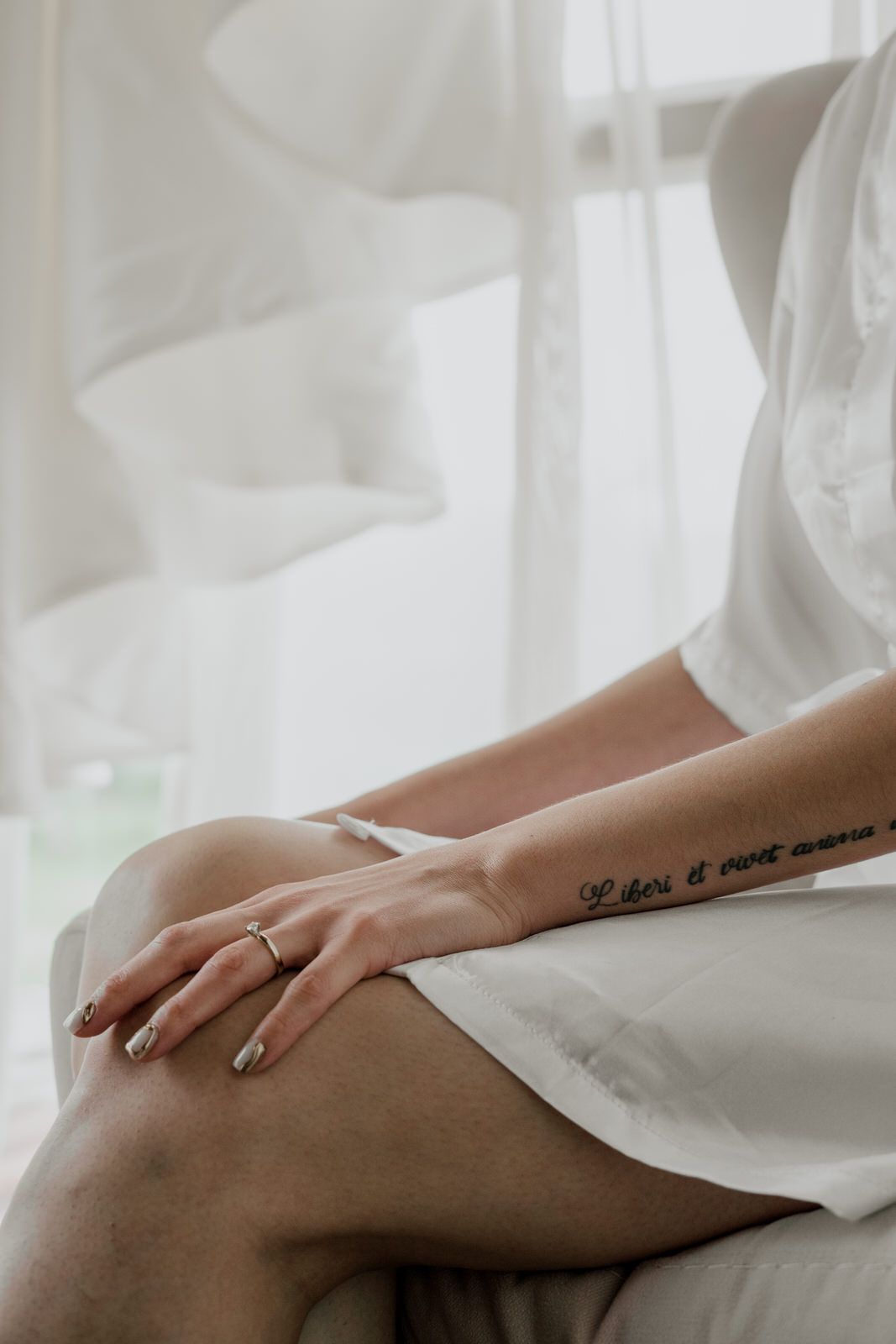 Close-up of hands with ring and tattoo against white fabric in soft window light.