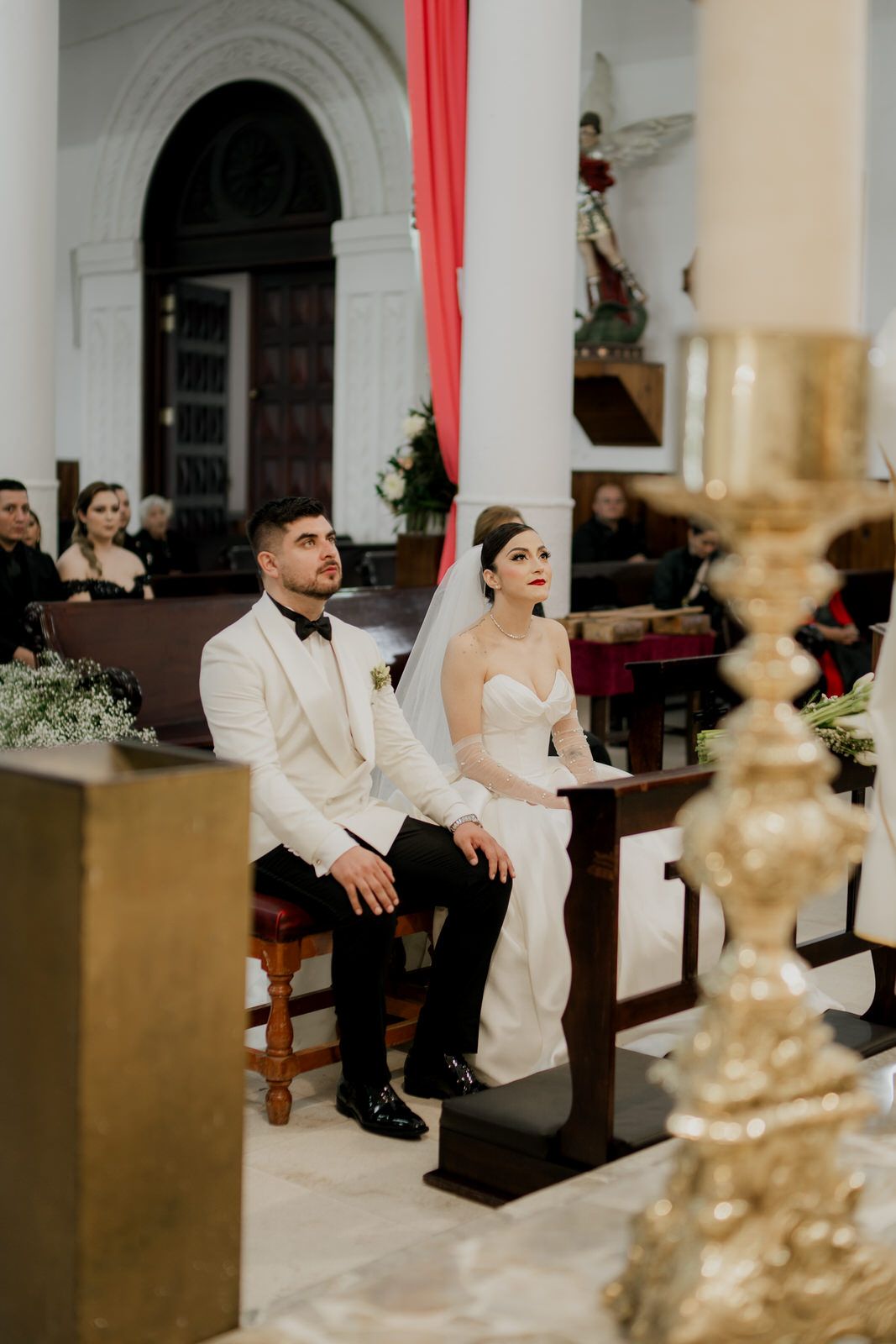 A couple seated during their wedding ceremony with elegant church architecture and decor visible.
