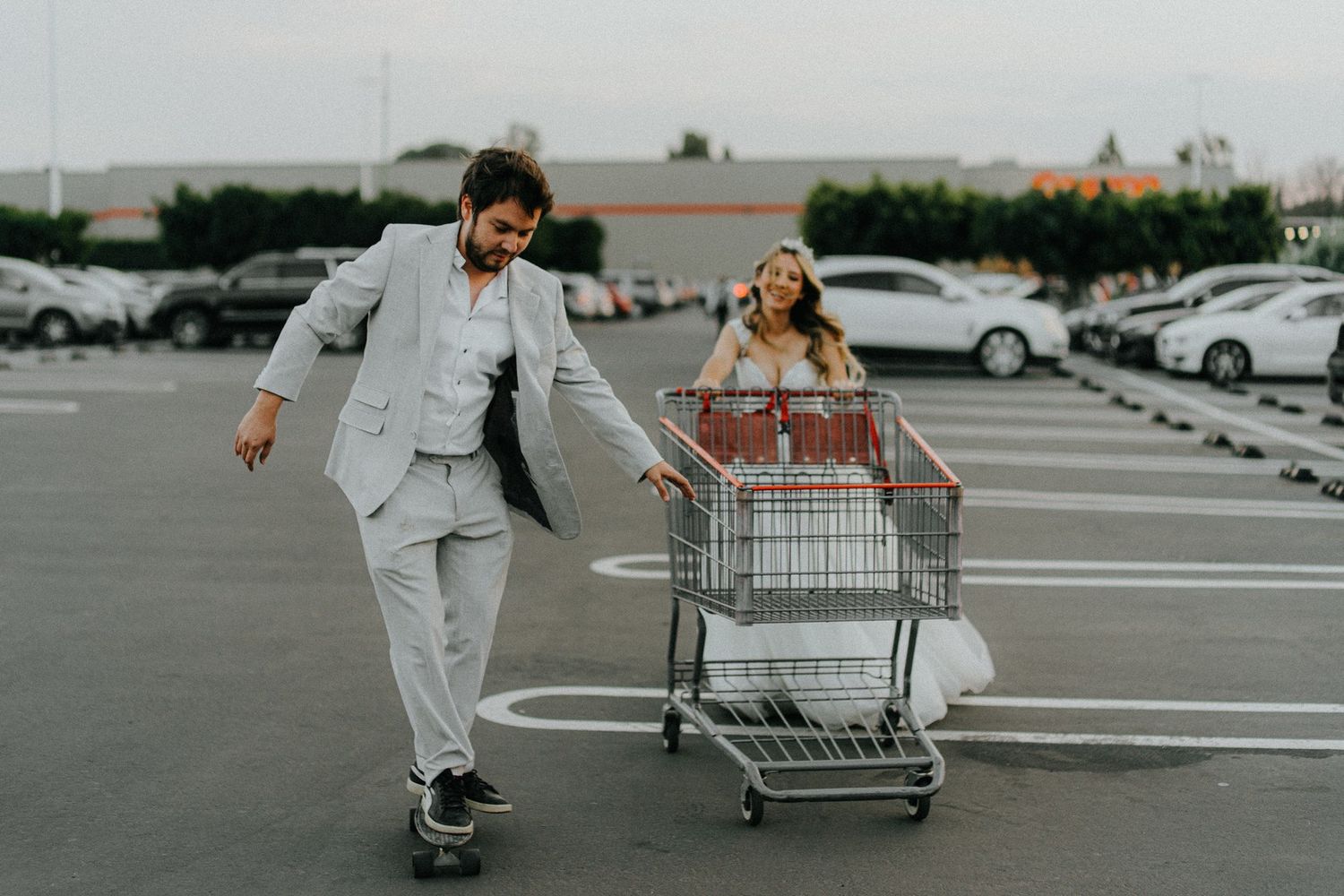 Couple in formal attire playfully pushing shopping cart in parking lot.