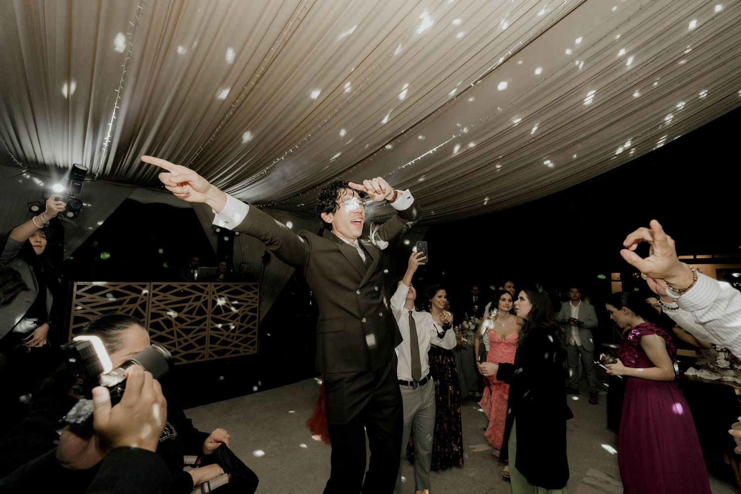 Energetic crowd dancing and jumping during a wedding celebration under a draped ceiling with twinkle lights.