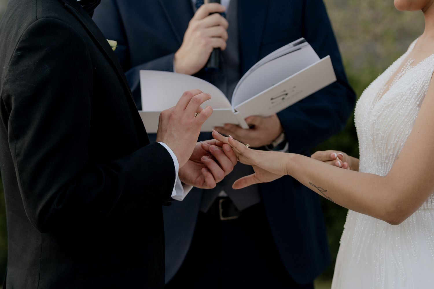 Groom placing wedding ring on bride's finger during outdoor ceremony while officiant holds booklet.