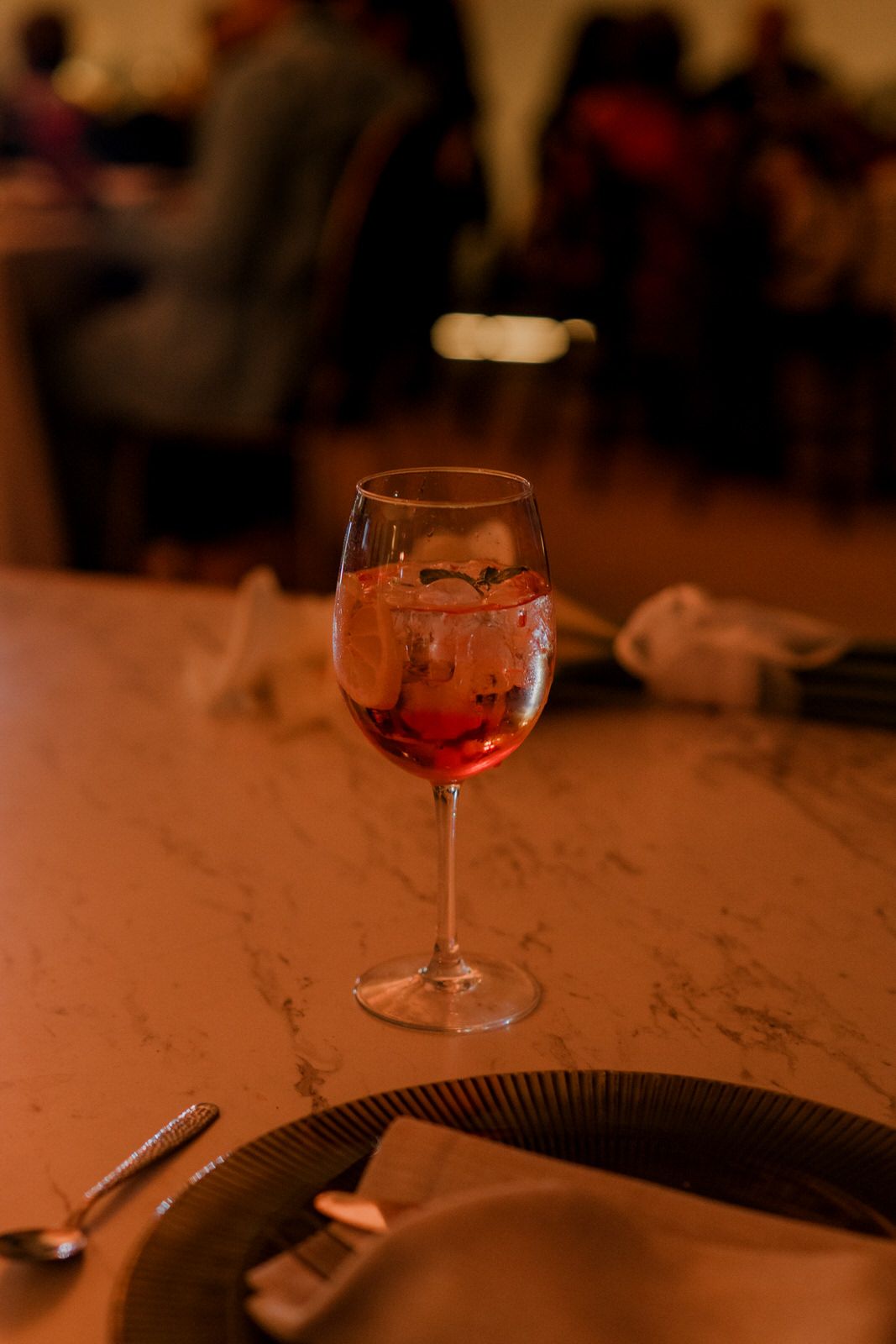 A red cocktail with ice in a wine glass on a marble table at a dimly lit restaurant.