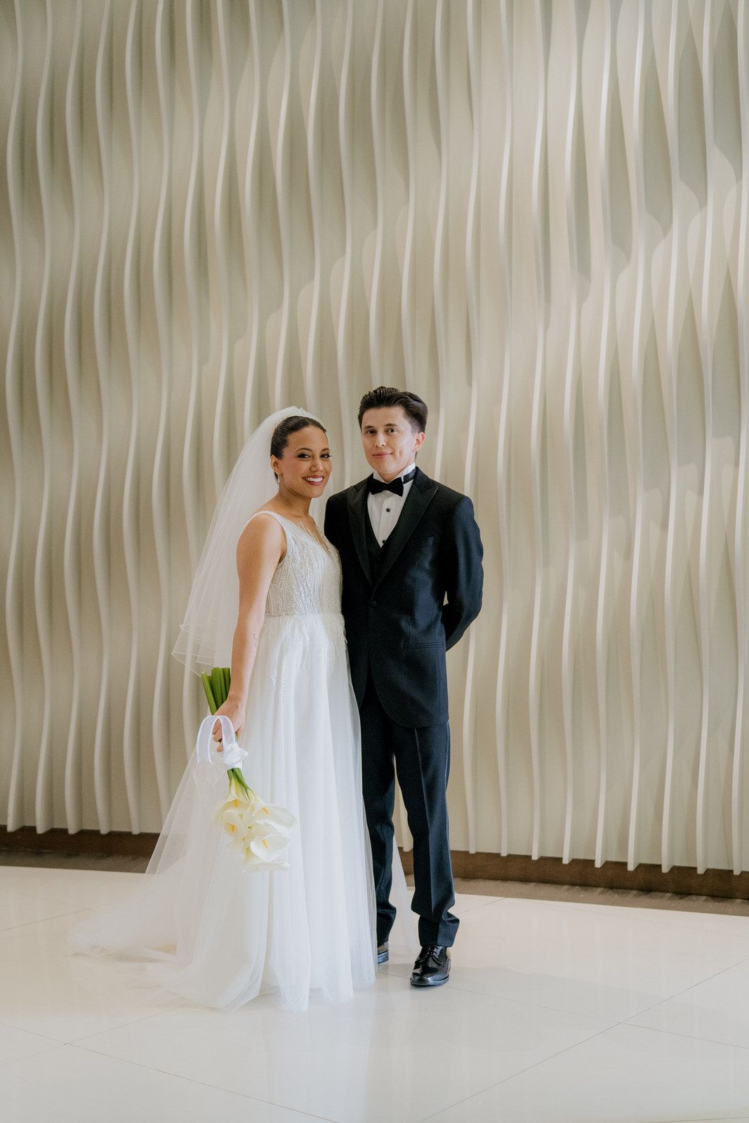 Bride in white gown and groom in black tuxedo posing together on their wedding day against a textured wall.