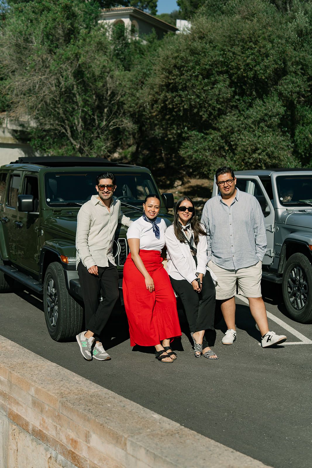A group poses together in front of a black Jeep in a sunny parking lot with trees in the background.