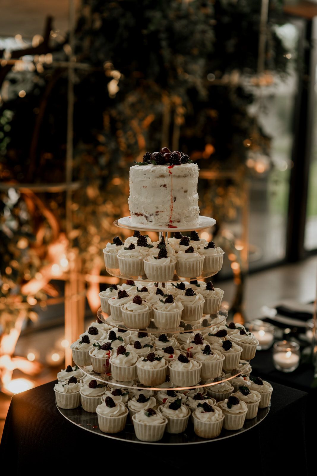 Tiered display of white cupcakes and cake decorated with dark berries at a wedding reception.