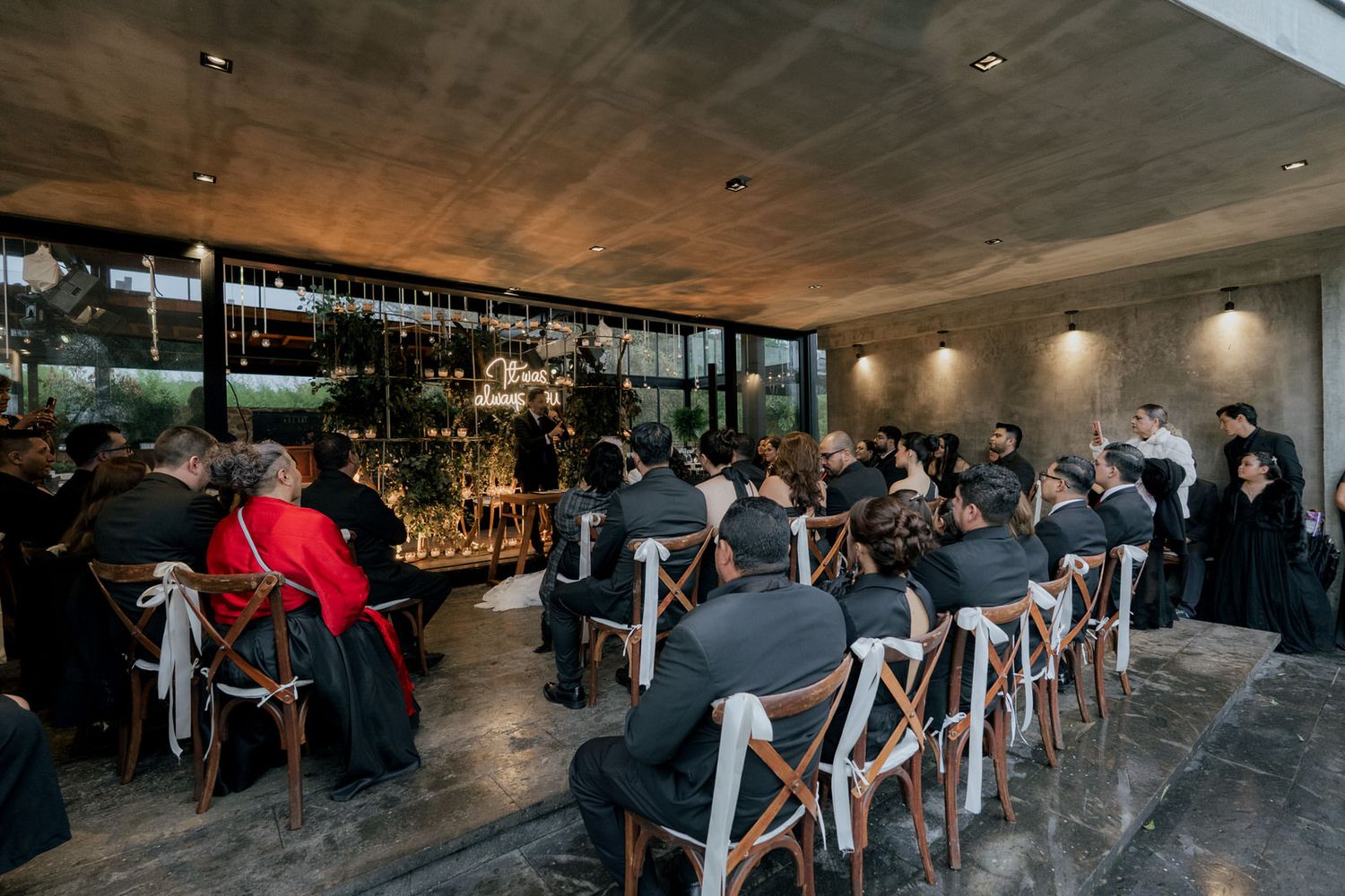 Wedding guests seated in modern venue with concrete ceiling and cross-back chairs.