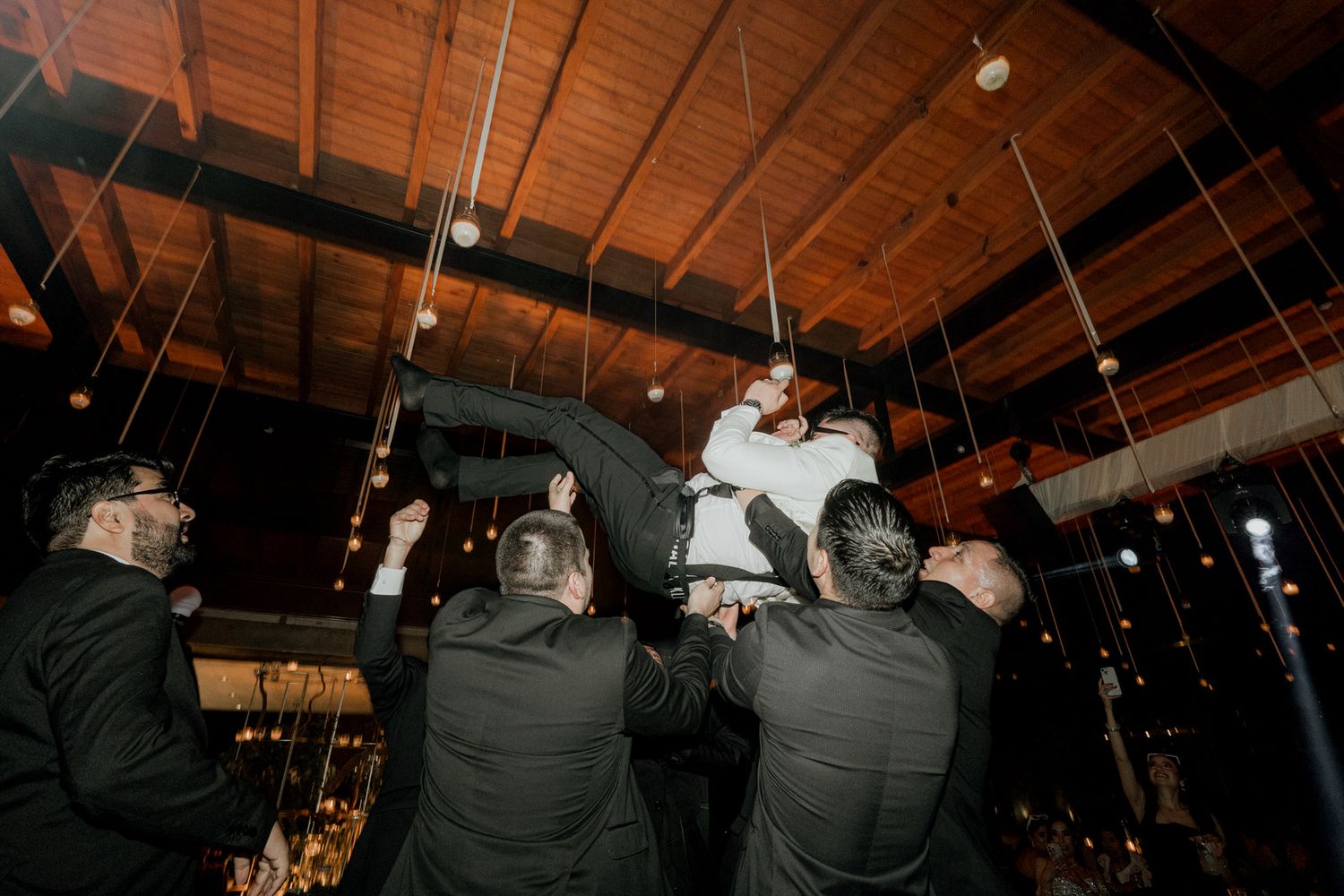 Wedding guests dancing together on a dark dance floor with wooden ceiling beams and hanging lights above.