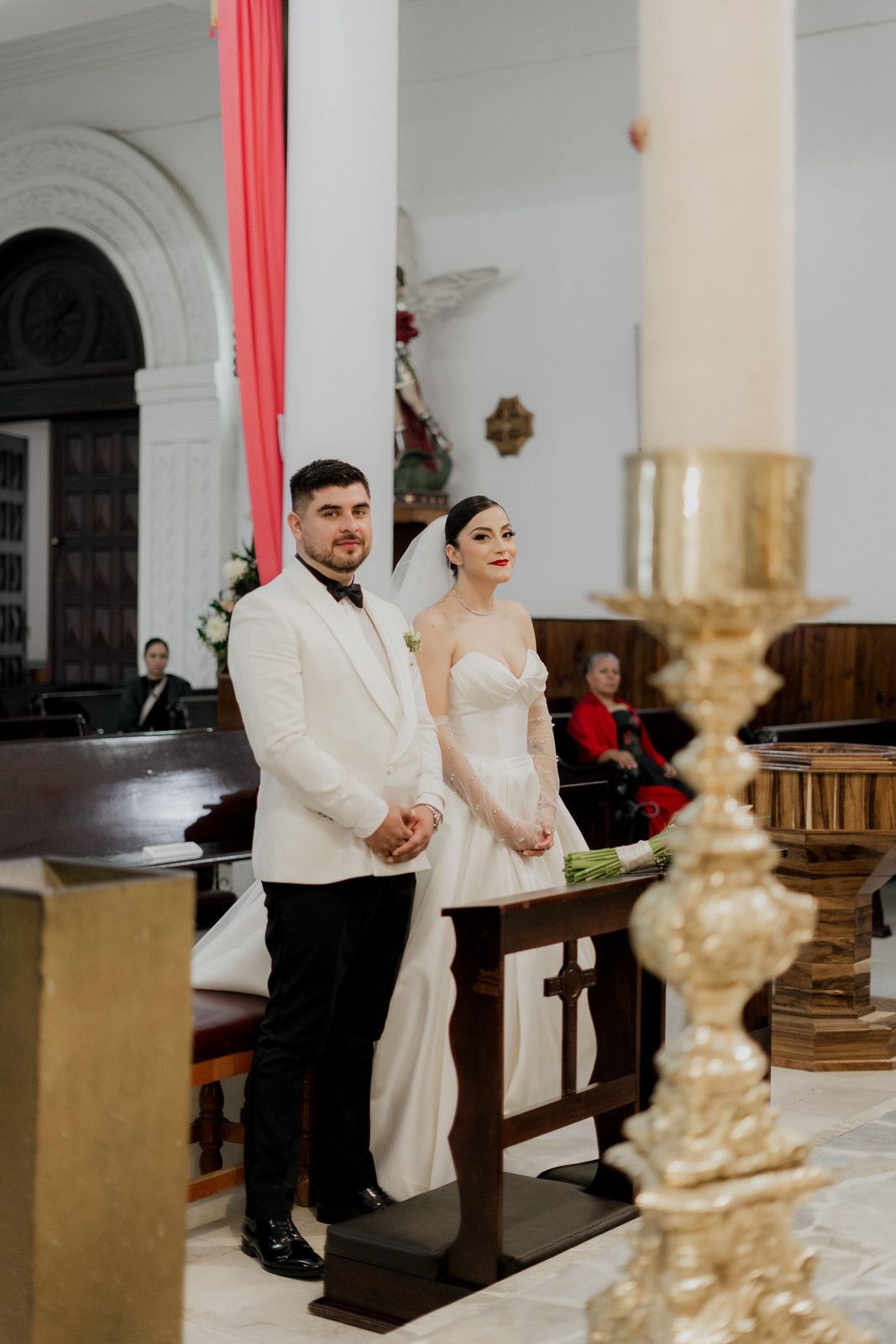 A couple stands at the altar during their wedding ceremony in a church with elegant white decor and red draping.