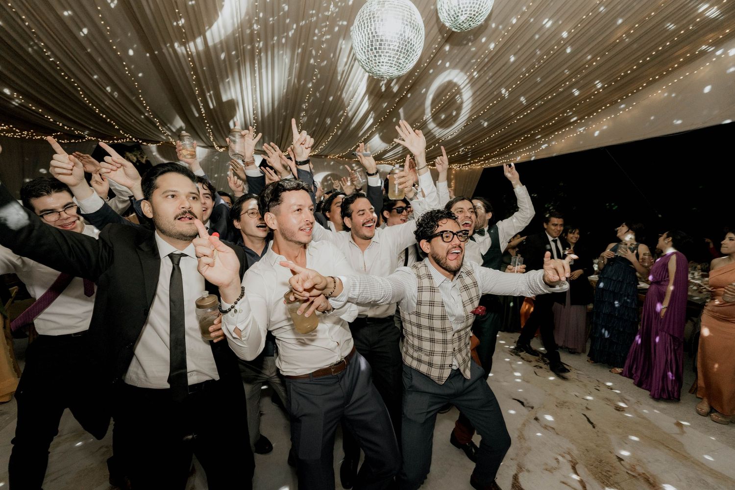Lively dance floor scene with guests celebrating under decorative lighting at an indoor wedding reception.