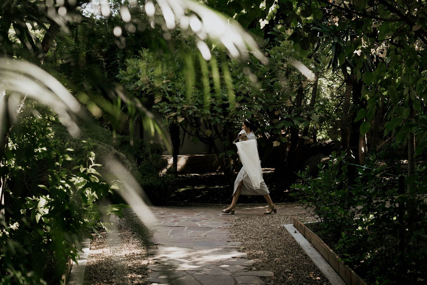 Figure in white dress walking along garden path surrounded by lush greenery.