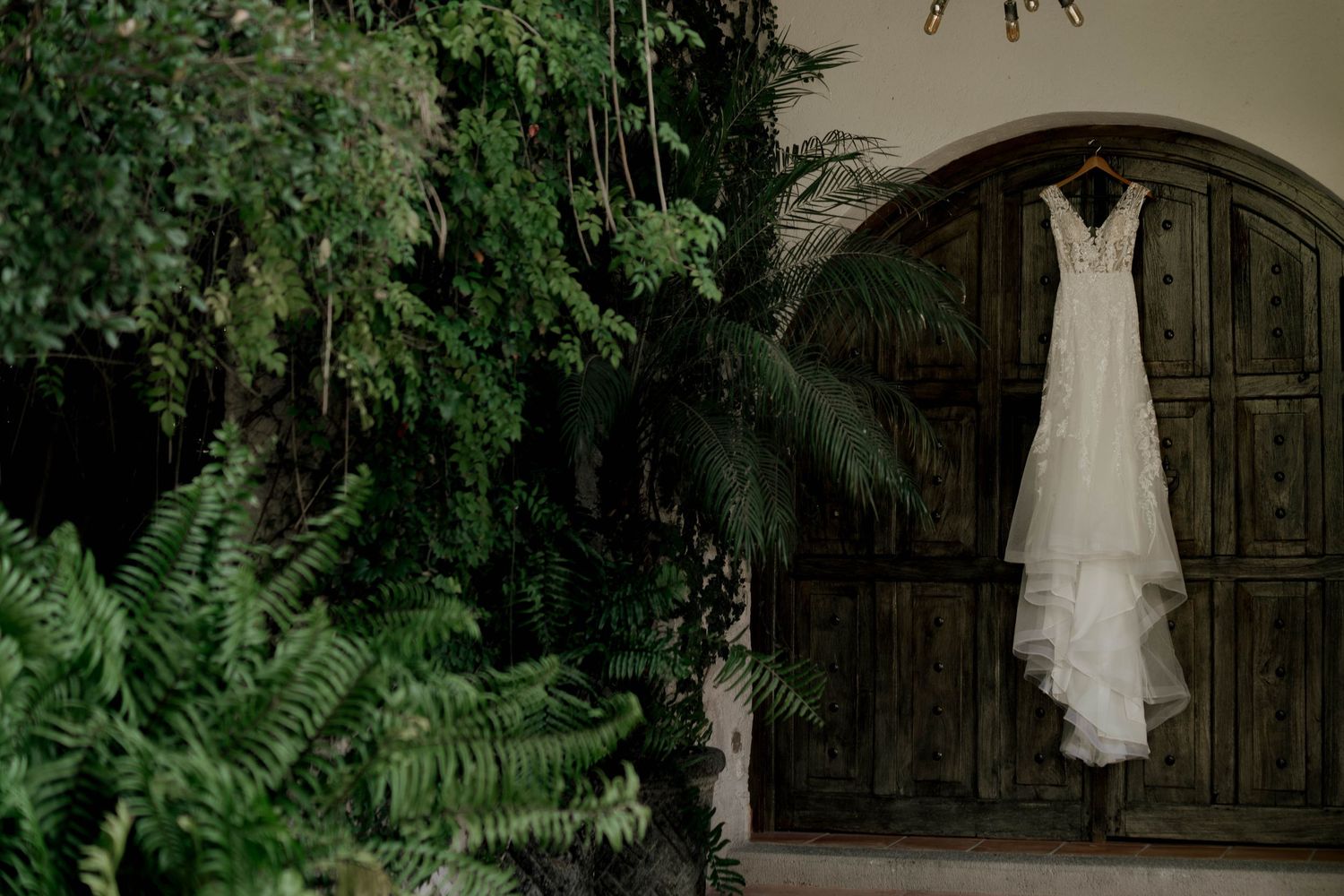 A beautiful white wedding dress hangs elegantly against a rustic wooden backdrop with green foliage.