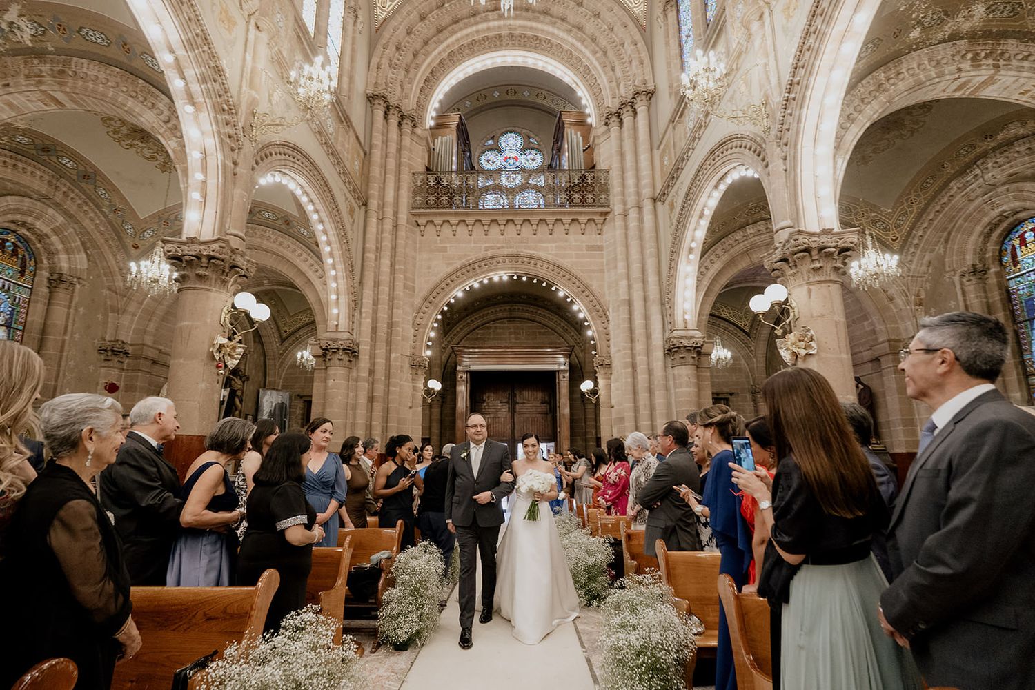 Bride walks down the aisle with escort in an ornate cathedral filled with wedding guests and floral decor.