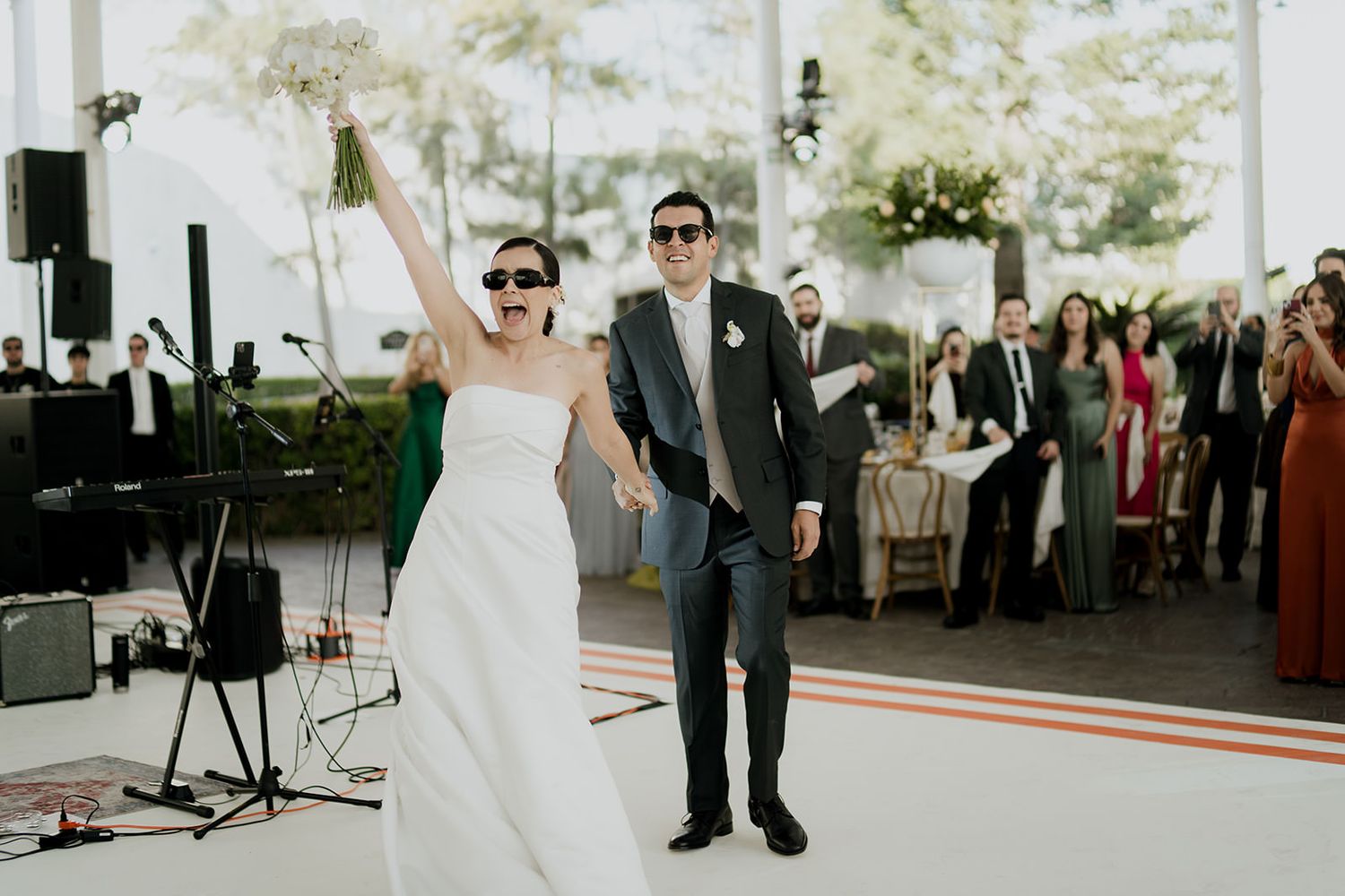 Newlyweds enter their wedding reception wearing sunglasses, bride raising bouquet under a white tent.