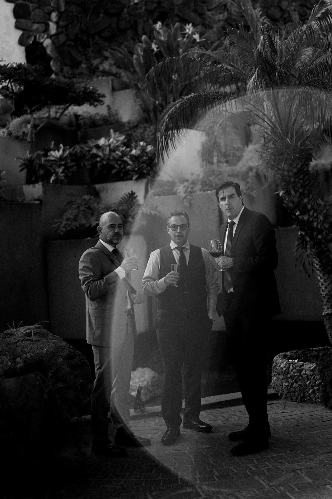 Black and white photo of three men in suits socializing outdoors at a wedding reception.