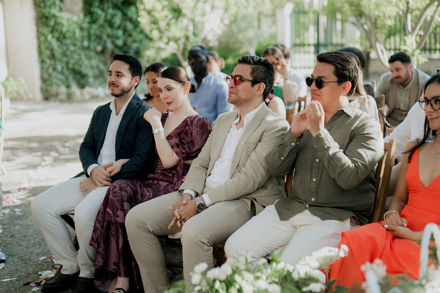 Wedding guests seated outdoors in rows smile and laugh during a ceremony in a garden setting.