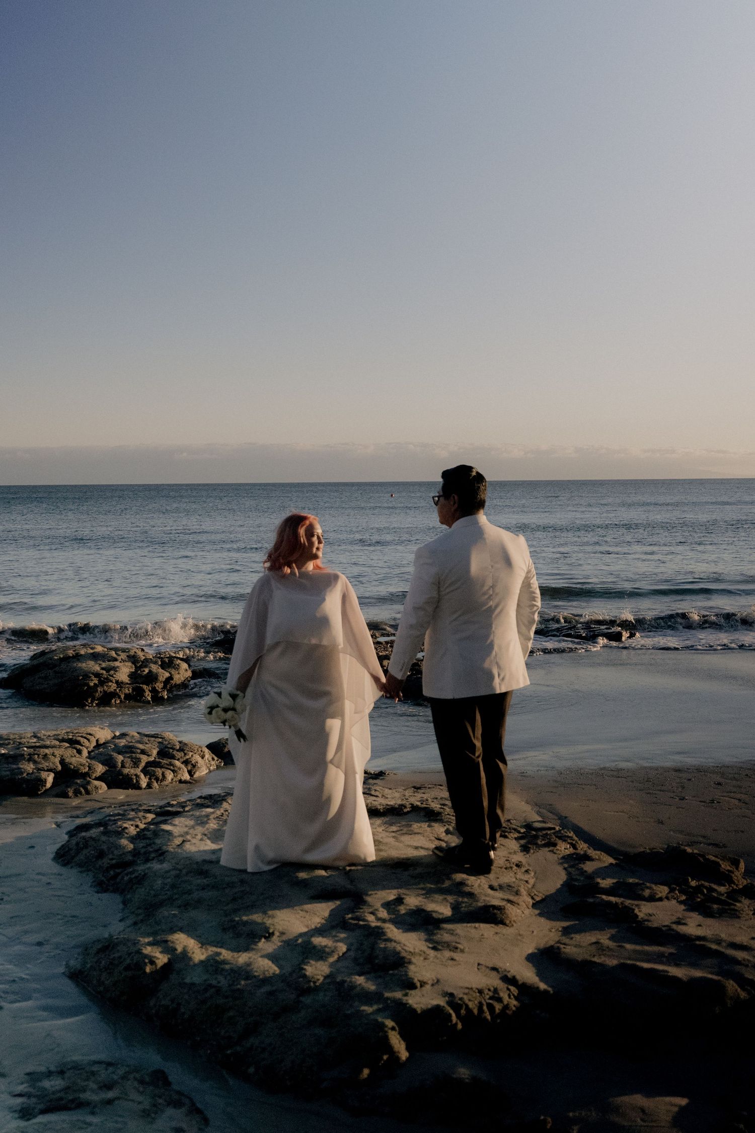 Bride and groom holding hands on rocky beach at sunset, wearing white wedding attire with ocean backdrop.