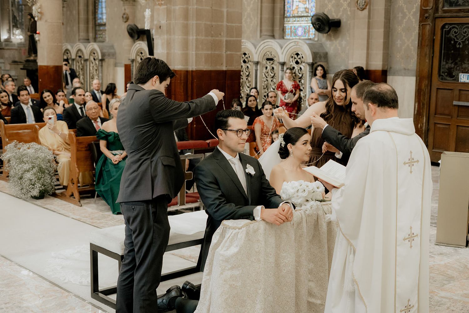 Couple kneeling at church altar during Catholic wedding ceremony with priest and guests in ornate cathedral.