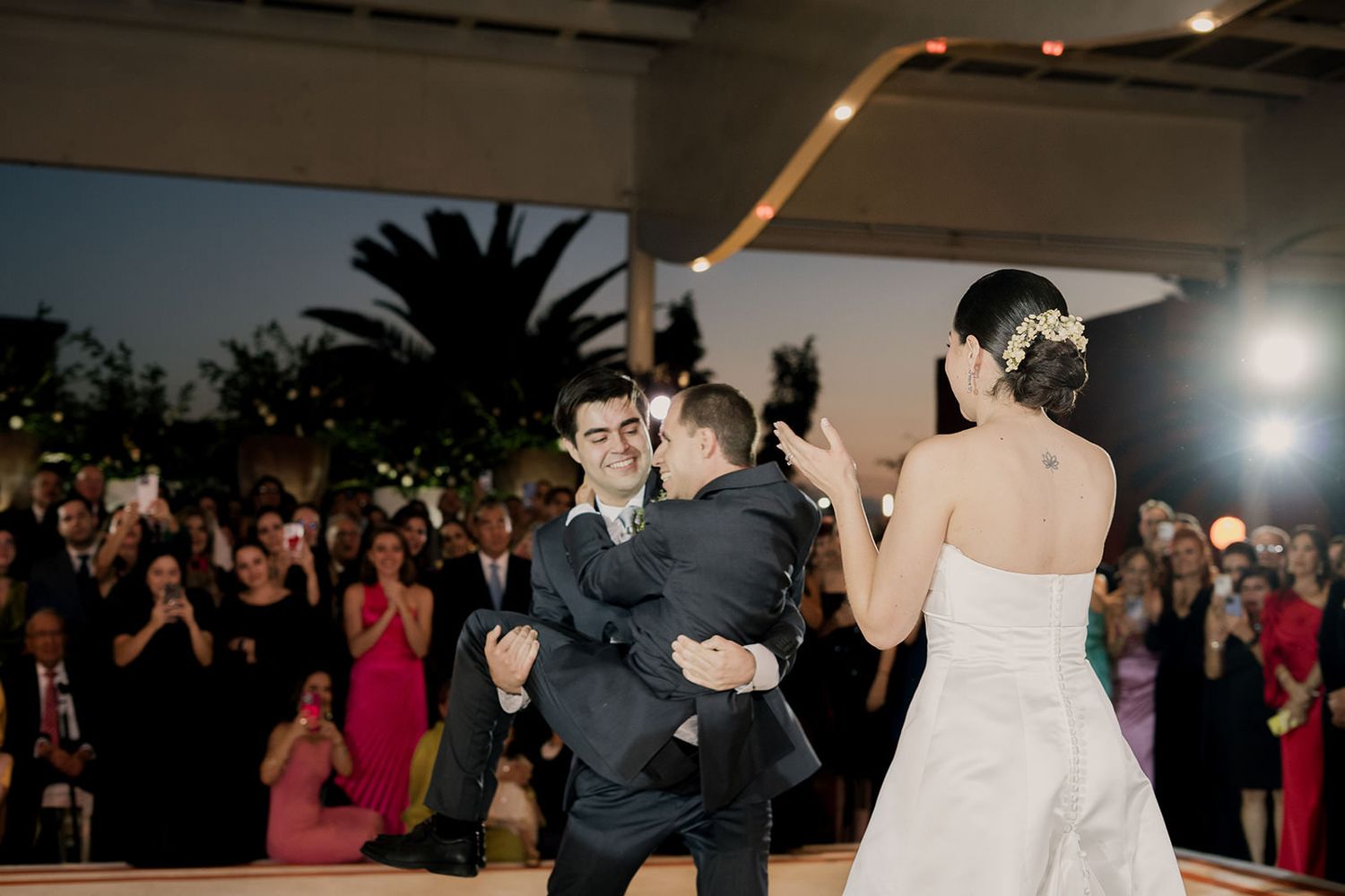 Groom embracing a guest during wedding first dance while bride watches at outdoor evening reception.