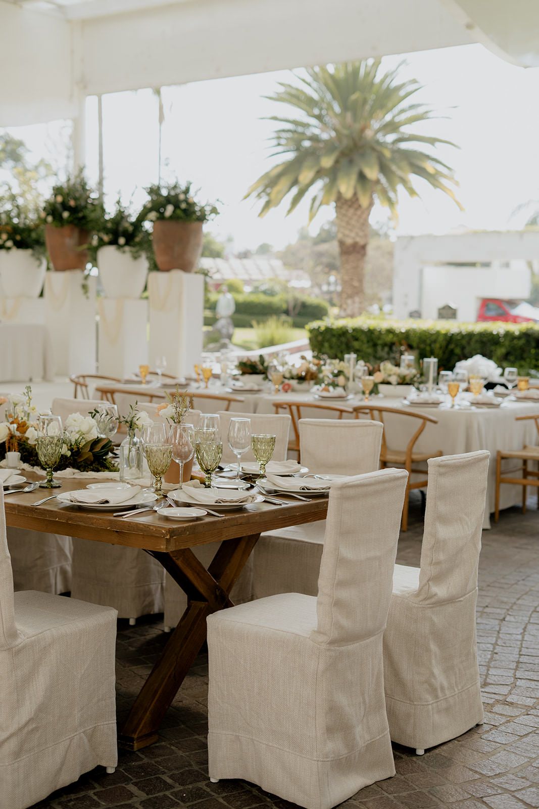 Elegant outdoor wedding reception with white linen chairs, rustic wood tables, floral centerpieces, and a palm tree backdrop.